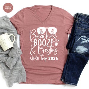 May include: A dusty rose V-neck t-shirt with the words "Beaches, Booze & Besties" in white script, along with a graphic of sunglasses, palm trees, and a cocktail. The text "Girls Trip 2026" is below the main text. The shirt is paired with blue jeans.