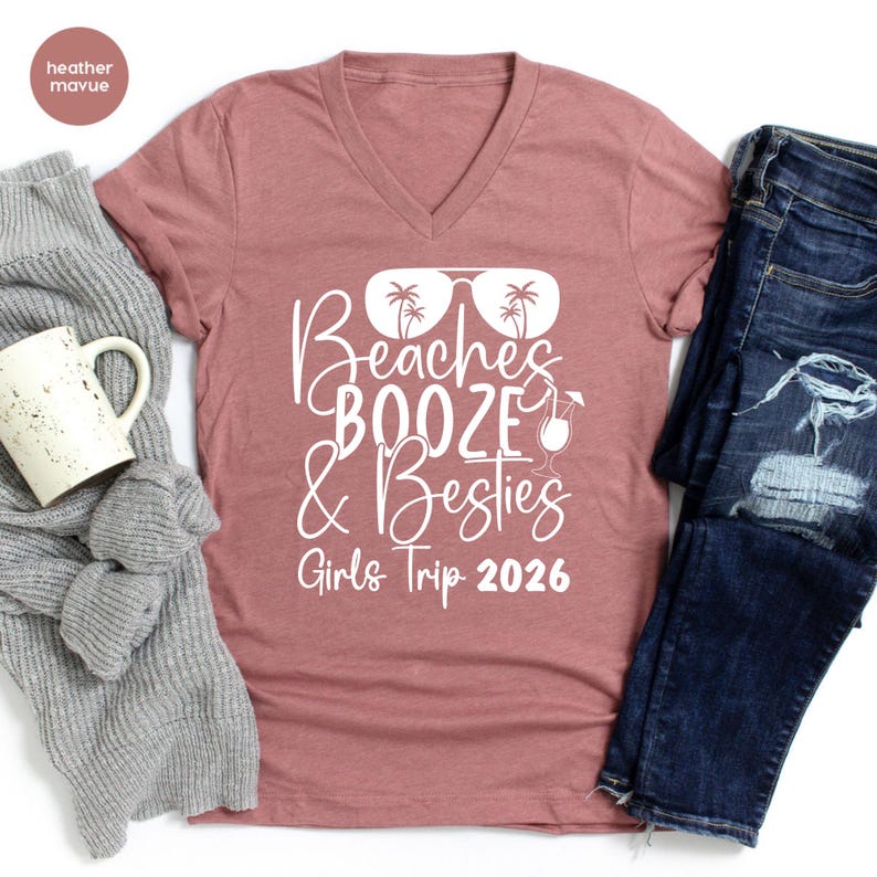 May include: A dusty rose V-neck t-shirt with the words "Beaches, Booze & Besties" in white script, along with a graphic of sunglasses, palm trees, and a cocktail. The text "Girls Trip 2026" is below the main text. The shirt is paired with blue jeans.