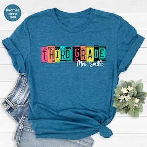 Op de afbeelding: Een heather deep teal t-shirt met korte mouwen en een afbeelding van kleurrijke nummerplaten die THIRD GRADE spellen in zwarte letters, met de tekst Mrs. Smith in wit cursief schrift onder de afbeelding van de nummerplaten.