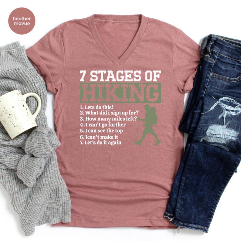 May include: A heather mauve V-neck t-shirt featuring white and green text that reads 7 STAGES OF HIKING followed by a numbered list of seven phrases and a silhouette of a hiker with a backpack and walking stick, alongside a gray sweater and jeans.