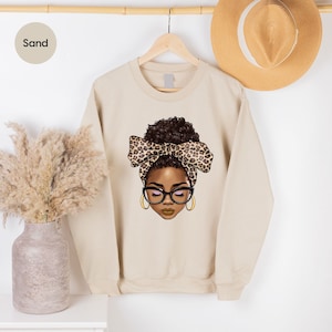 May include: A sand-colored sweatshirt with a graphic of a woman with brown skin, brown hair, and a leopard print headband. The woman is wearing glasses and has a brown bow in her hair.
