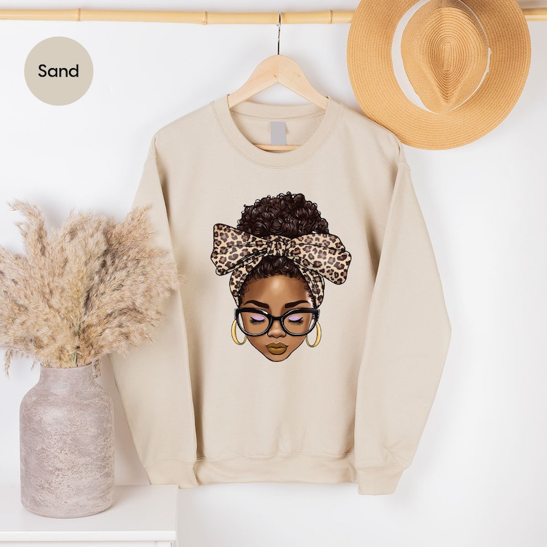 May include: A sand-colored sweatshirt with a graphic of a woman with brown skin, brown hair, and a leopard print headband. The woman is wearing glasses and has a brown bow in her hair.