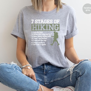 May include: A heather grey short-sleeve t-shirt featuring white and olive green text that reads 7 STAGES OF HIKING followed by a numbered list of seven phrases and a silhouette graphic of a hiker with a backpack and a walking stick.