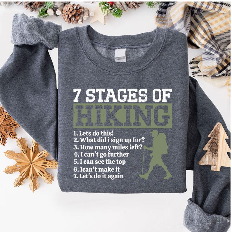 May include: A dark gray crewneck sweatshirt featuring white and olive green text that reads 7 STAGES OF HIKING followed by a numbered list of seven phrases and a silhouette of a hiker with a backpack and walking stick on the right side.