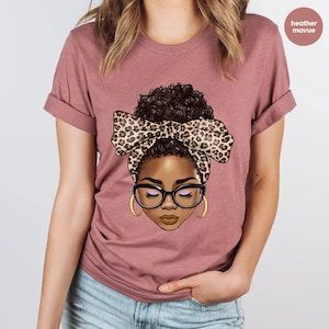 May include: A heather mauve t-shirt with a graphic of a woman with brown hair in a bun wearing a leopard print headband and glasses.