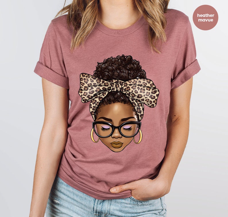 May include: A heather mauve t-shirt with a graphic of a woman with brown hair in a bun wearing a leopard print headband and glasses.