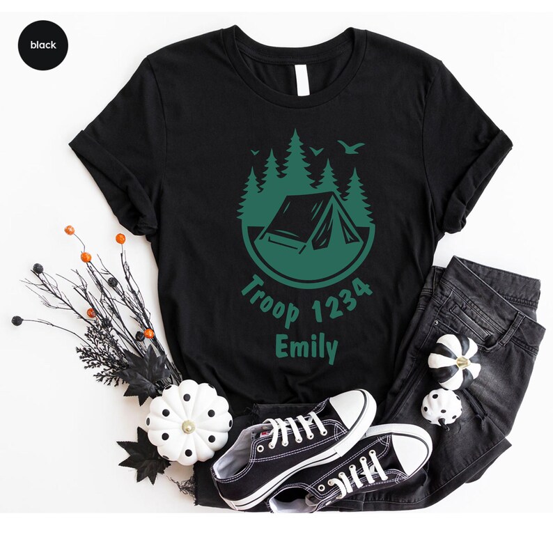 Może przedstawiać: Czarny t-shirt z zieloną grafiką kempingową w okręgu. Tekst "Troop 1234 Emily" znajduje się poniżej. Obraz zawiera czarne dżinsy i czarno-białe trampki.