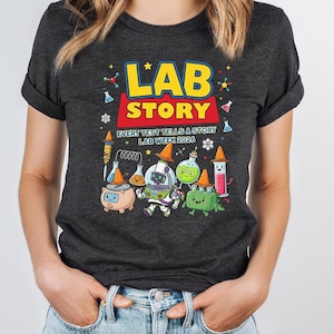 Op de afbeelding: Donkergrijs t-shirt met een kleurrijke afbeelding met de tekst "LAB STORY" in geel en rood, met de tekst "EVERY TEST TELLS A STORY LAB WEEK 2026". De afbeelding toont cartoon laboratoriumapparatuur en -personages.