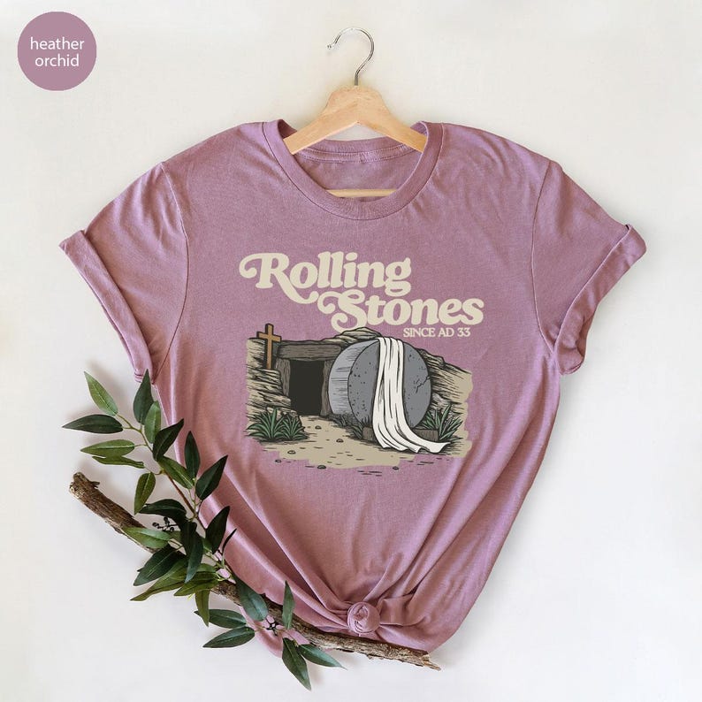 May include: A heather orchid t-shirt with a graphic of a stone tomb with a white sheet draped over it. The text "Rolling Stones Since AD 33" is above the tomb.