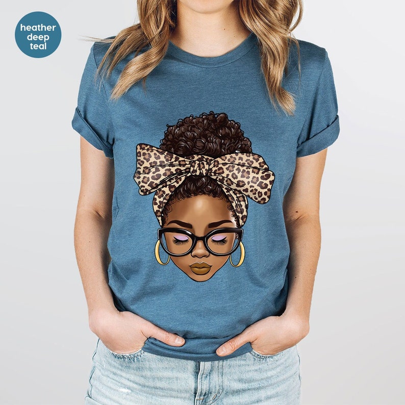 May include: A blue t-shirt with a graphic of a woman with brown skin, brown hair in a bun, wearing glasses and a leopard print headband.