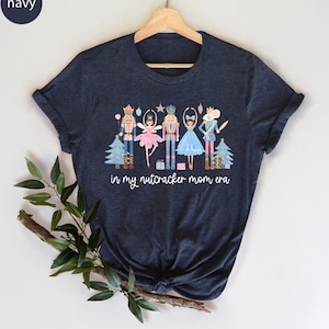 May include: Navy blue t-shirt featuring a whimsical Nutcracker-themed design. The graphic includes illustrations of Nutcracker characters, Christmas trees, and the text "in my nutcracker mom era". A festive and fun tee.