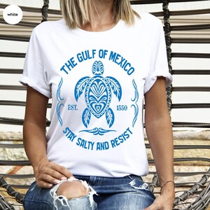 Puede incluir: Camiseta blanca con un gr&aacute;fico azul de una tortuga y el texto "The Gulf of Mexico Est. 1550 Stay Salty and Resist".