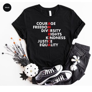 May include: Black t-shirt with the word "AMERICA" spelled vertically with the letters in red. The words "COURAGE, FREEDOM, DIVERSITY, RIGHTS, KINDNESS, JUSTICE, EQUALITY" are printed in white. The shirt is paired with black jeans and black sneakers.