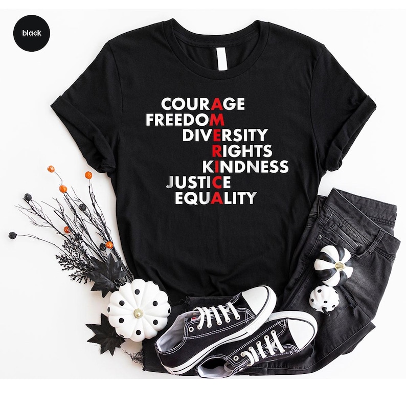 May include: Black t-shirt with the word "AMERICA" spelled vertically with the letters in red. The words "COURAGE, FREEDOM, DIVERSITY, RIGHTS, KINDNESS, JUSTICE, EQUALITY" are printed in white. The shirt is paired with black jeans and black sneakers.