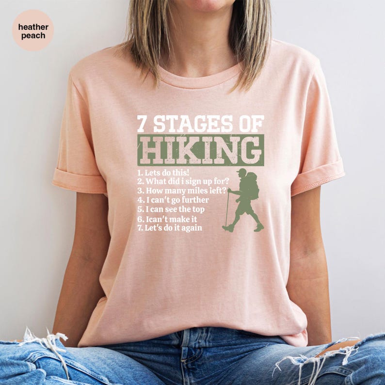 May include: A heather peach t-shirt featuring the text 7 STAGES OF HIKING in white and green, followed by a numbered list of seven phrases and a silhouette of a hiker with a backpack and walking stick, all printed in a distressed white font.