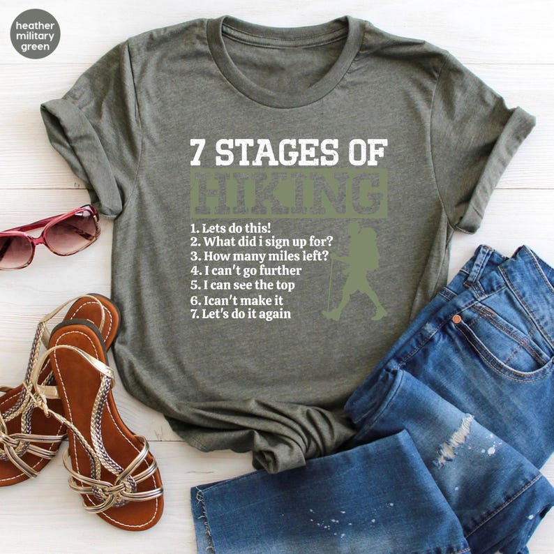 May include: A heather military green t-shirt featuring the white text 7 STAGES OF HIKING and a list of seven numbered phrases about hiking, accompanied by a silhouette of a hiker with a backpack and a walking stick on the front center.