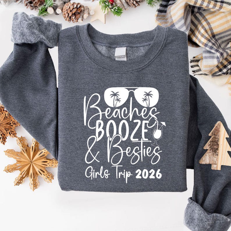 May include: A dark gray sweatshirt with the words "Beaches, Booze & Besties, Girls Trip 2026" in white script. The design includes sunglasses with palm trees and a cocktail glass. The sweatshirt has a crew neck and long sleeves.