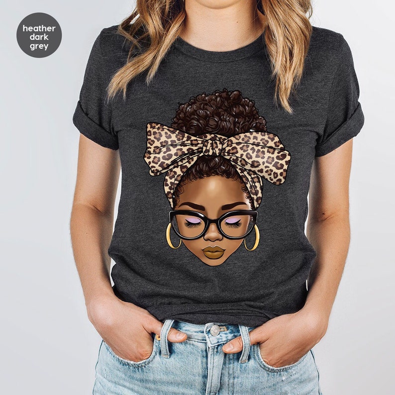 May include: A heather dark grey t-shirt with a graphic of a woman with brown hair in a bun wearing a leopard print headband and glasses.