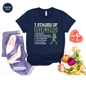 May include: A navy blue t-shirt featuring the white text 7 STAGES OF HIKING and a list of seven hiking-related phrases alongside a silhouette of a hiker with a backpack and walking stick, paired with blue denim jeans and white high heels.
