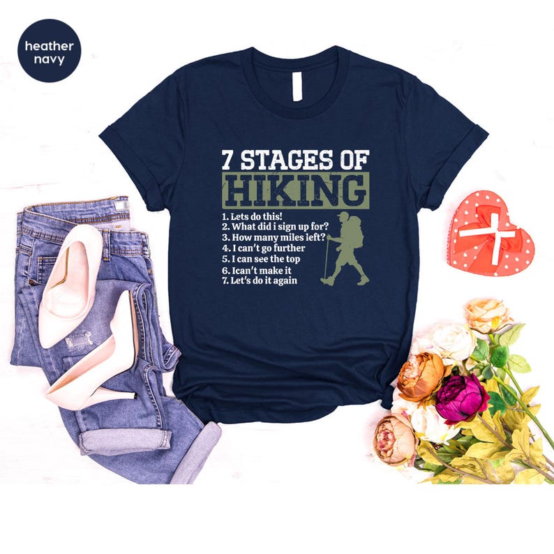 May include: A navy blue t-shirt featuring the white text 7 STAGES OF HIKING and a list of seven hiking-related phrases alongside a silhouette of a hiker with a backpack and walking stick, paired with blue denim jeans and white high heels.
