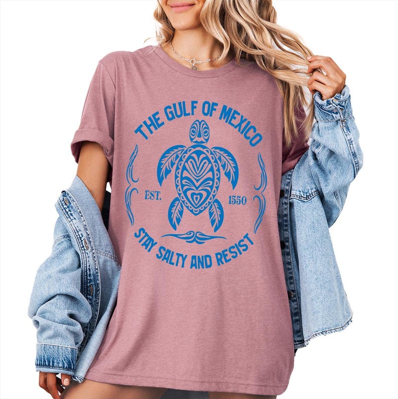 Puede incluir: Una camiseta rosa con un gr&aacute;fico azul de una tortuga con el texto "The Gulf of Mexico Est. 1550 Stay Salty and Resist".