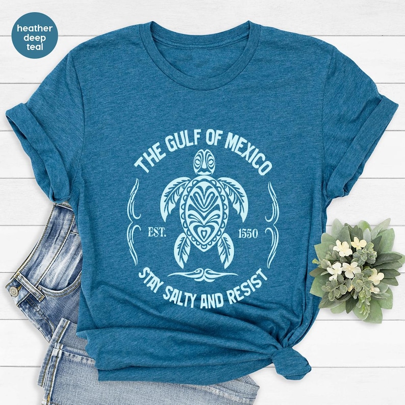 Puede incluir: Una camiseta azul turquesa con un dise&ntilde;o gr&aacute;fico blanco de una tortuga marina con el texto "The Gulf of Mexico Est. 1550 Stay Salty and Resist".