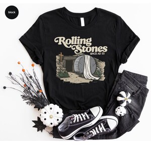 May include: Black t-shirt with a graphic of a stone tomb with a white sheet on top. The text "Rolling Stones Since AD 33" is above the tomb.