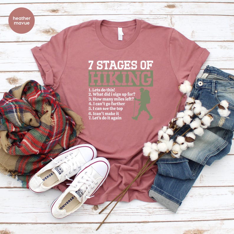 May include: A heather mauve t-shirt featuring the white text 7 STAGES OF HIKING with a green rectangular background behind the word HIKING and a list of seven hiking-related phrases alongside a green silhouette of a hiker with a backpack.