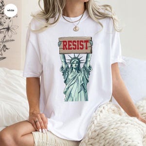 May include: White t-shirt with a graphic of the Statue of Liberty holding a sign that reads "RESIST" in red. The Statue of Liberty is illustrated in green and blue tones.