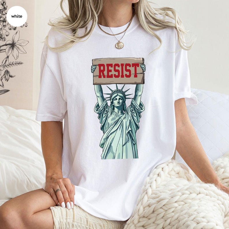 May include: White t-shirt with a graphic of the Statue of Liberty holding a sign that reads "RESIST" in red. The Statue of Liberty is illustrated in green and blue tones.