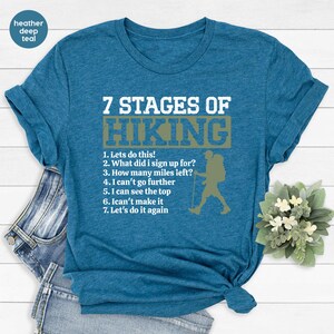 May include: A heather deep teal short-sleeve t-shirt featuring white text that reads 7 STAGES OF HIKING followed by a numbered list of seven stages and a graphic silhouette of a hiker with a backpack and walking stick in a muted green box.