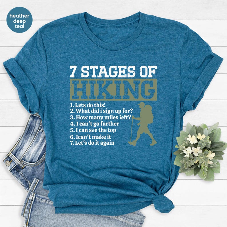 May include: A heather deep teal short-sleeve t-shirt featuring white text that reads 7 STAGES OF HIKING followed by a numbered list of seven stages and a graphic silhouette of a hiker with a backpack and walking stick in a muted green box.