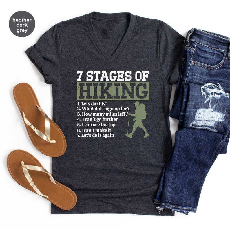 May include: A heather dark grey V-neck t-shirt featuring the white text 7 STAGES OF HIKING, a list of seven numbered phrases, and a silhouette of a hiker with a backpack and walking stick, accompanied by brown flip-flops and blue denim jeans.
