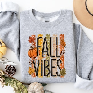 May include: A heather gray crewneck sweatshirt featuring a graphic print with the text FALL VIBES in black. The design includes vertical stripes in orange, yellow, and green with white polka dots, accented by orange pumpkin and leaf illustrations.