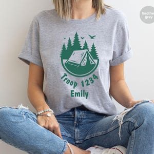 Może przedstawiać: Szara koszulka z nadrukiem w kolorze zielonym przedstawiającym namiot, drzewa i napis "Troop 1234 Emily". Koszulka wykonana jest z miękkiego materiału i ma kr&oacute;tkie rękawy.