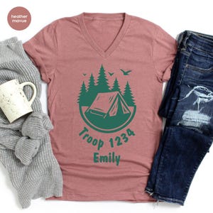 Może przedstawiać: R&oacute;żowa koszulka z dekoltem w serek z zieloną grafiką kempingową. Grafika zawiera namiot, drzewa i tekst "Troop 1234 Emily". Koszulka jest połączona z szarym swetrem, białym kubkiem i niebieskimi dżinsami.