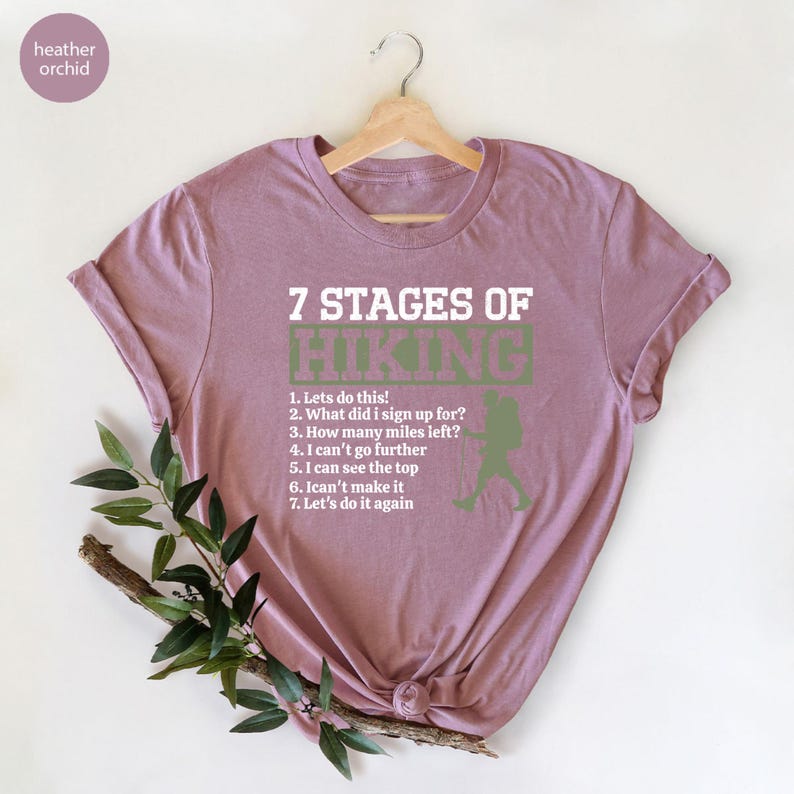 May include: A heather orchid short-sleeve t-shirt featuring white text that reads 7 STAGES OF HIKING followed by a numbered list of seven phrases and a green silhouette of a hiker with a backpack, all printed on the front of the garment.