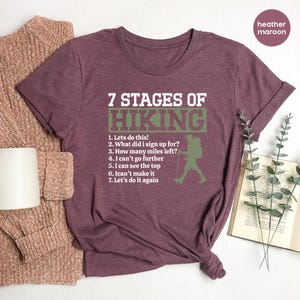 May include: A heather maroon short-sleeve t-shirt featuring the white text 7 STAGES OF HIKING above a list of seven numbered phrases and a green silhouette of a hiker with a backpack and trekking pole, all printed on the front of the garment.