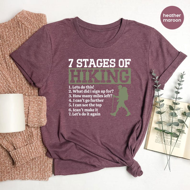 May include: A heather maroon short-sleeve t-shirt featuring the white text 7 STAGES OF HIKING above a list of seven numbered phrases and a green silhouette of a hiker with a backpack and trekking pole, all printed on the front of the garment.
