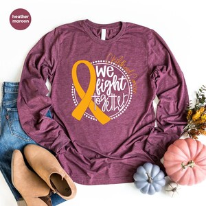 May include: A heather maroon long-sleeve t-shirt with a gold ribbon and the text "Childhood Cancer We fight together" in a white circle.