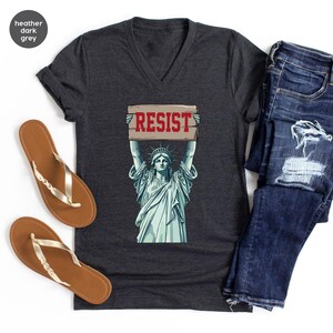 May include: Heather dark grey V-neck t-shirt with a graphic of the Statue of Liberty holding a sign with the word "RESIST" in red. The shirt is styled with blue jeans and brown sandals.