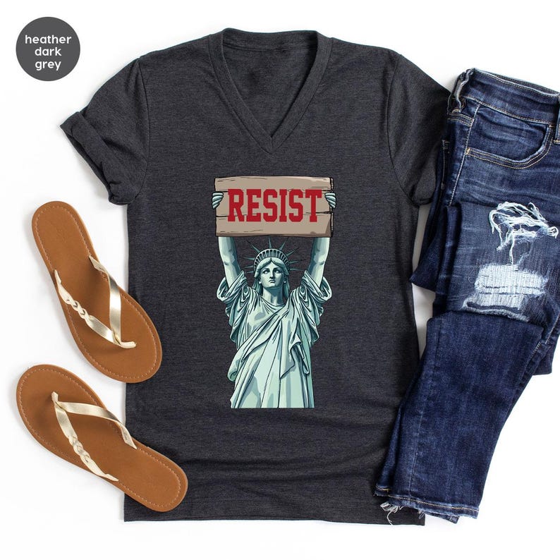 May include: Heather dark grey V-neck t-shirt with a graphic of the Statue of Liberty holding a sign with the word "RESIST" in red. The shirt is styled with blue jeans and brown sandals.