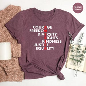 May include: Heather maroon t-shirt with the words COURAGE, FREEDOM, DIVERSITY, RIGHTS, KINDNESS, JUSTICE, and EQUALITY printed in white and red. A folded sweater and a white mug are on the left. A book and sprigs of greenery are on the right.