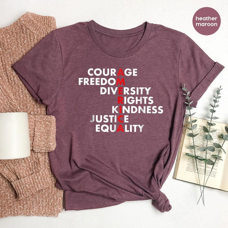 May include: Heather maroon t-shirt with the words COURAGE, FREEDOM, DIVERSITY, RIGHTS, KINDNESS, JUSTICE, and EQUALITY printed in white and red. A folded sweater and a white mug are on the left. A book and sprigs of greenery are on the right.