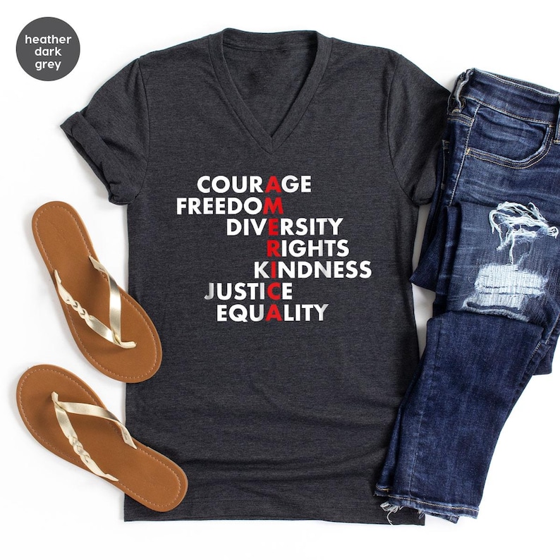 May include: Heather dark grey V-neck t-shirt with the word "AMERICA" spelled vertically with the words "Courage, Freedom, Diversity, Rights, Kindness, Justice, Equality". The shirt is paired with blue jeans and brown sandals.