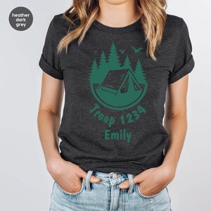 Może przedstawiać: Ciemnoszara koszulka z nadrukiem w kolorze zielonym przedstawiającym namiot, drzewa i ptaki, z napisem "Troop 1234 Emily". Koszulka ma podwinięte rękawy.