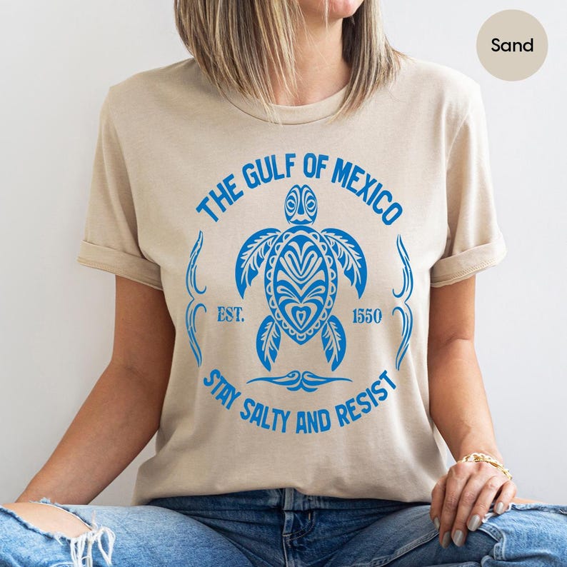 Puede incluir: Una camiseta beige con un gr&aacute;fico azul de una tortuga marina y el texto "The Gulf of Mexico Est. 1550 Stay Salty and Resist".