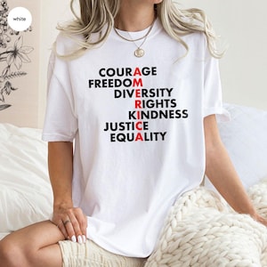 May include: White t-shirt with the word "AMERICA" spelled out vertically using words like "Courage", "Freedom", "Diversity", "Rights", "Kindness", "Justice", and "Equality". The letters "A", "R", and "A" are in red.