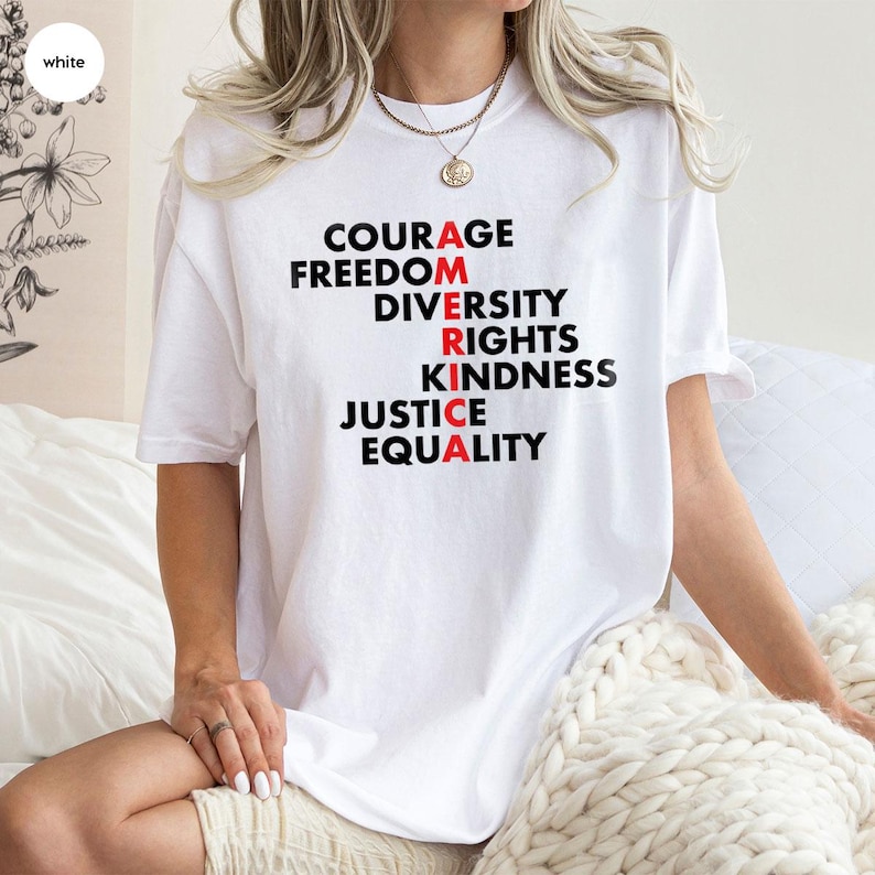 May include: White t-shirt with the word "AMERICA" spelled out vertically using words like "Courage", "Freedom", "Diversity", "Rights", "Kindness", "Justice", and "Equality". The letters "A", "R", and "A" are in red.