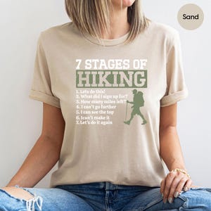 May include: A sand-colored t-shirt featuring the text 7 STAGES OF HIKING in white and green, followed by a numbered list of seven hiking-related phrases and a green silhouette of a hiker with a backpack and walking stick on the front.